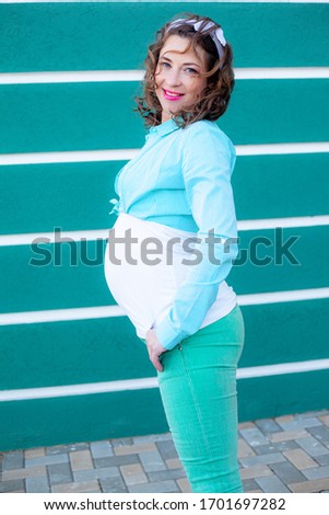 Happiness pregnant woman with  seven month tummy walking outdoor against the blue street wall. Female dressed white t-shirt , shirt and jeans