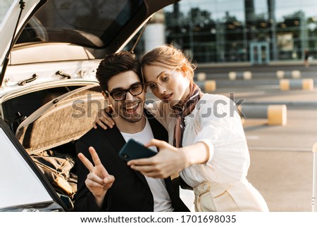 Cheerful brunette man in sunglasses shows v-sign. Blonde woman takes selfie with boyfriend. Guy in black suit sits in car and lady in eyeglasses holds phone.