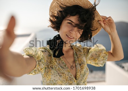 Brunette charming woman in boater takes selfie on sea background. Attractive lady in hat and floral outfit poses on balcony with ocean view.
