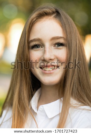 Emotional portrait of a tender and cheerful beautiful teenager girl in a white shirt, with a laugh, looking at her friend against the background of blurry foliage while walking in the city. Summer hot