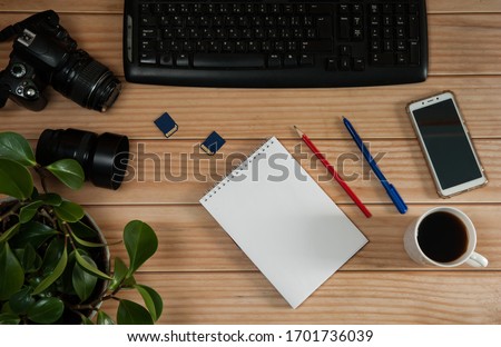 The concept of remote work at home. Camera, lens, black coffee mug, keyboard, blank notepad with pencil and pen close-up on a wooden background.
