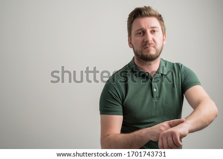 Portrait of serious stylish attractive man with thick beard, dressed in casual green t shirt showing late gesture isolated on gray background with copy space advertising area