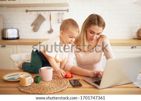 Beautiful young woman sitting in front of open laptop with her baby son on kitchen table watching cartoons online. Cute mother using electronic portable computer spending time at home with child
