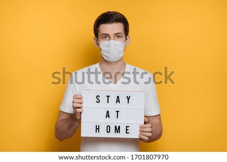 Young man in protective face mask holding lightbox with message to Stay Home to prevent spread of coronavirus infection. Covid-19 quarantine, self isolation. Isolated on yellow background