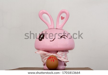 A adorable rabbit with a pink cover cloth is looking straight and egg that written Covid-19 is in front of it.