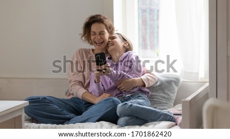 Cheerful mom and teen daughter laughing using smartphone app at home sitting on bed. Happy positive family adult parent having fun watching funny videos enjoying lifestyle relaxing with teenage girl. Royalty-Free Stock Photo #1701862420