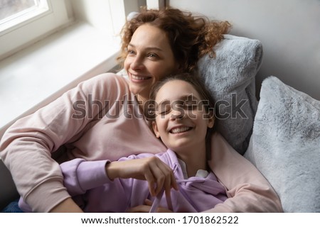 Relaxed affectionate family young adult single mom and cute teenage daughter resting hugging lying on bed together. Smiling mum embracing school kid girl bonding cuddling, lounge in bedroom, top view.