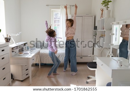 Carefree funny family young mum and teenage daughter dancing jumping together at home. Happy cool parent mother having fun with teen child girl enjoying leisure activity in cozy bedroom interior.