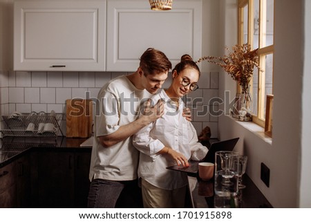 positive caucasian married couple use laptop at home, they spend morning together in the kitchen, look at screen of laptop, watch something interesting