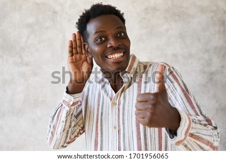 Isolated image of positive attractive young dark skinned guy making approval gesture, showing thumbs up sign, holding hand at his ear and smiling broadly, enjoying intriguing news, gossips and rumors