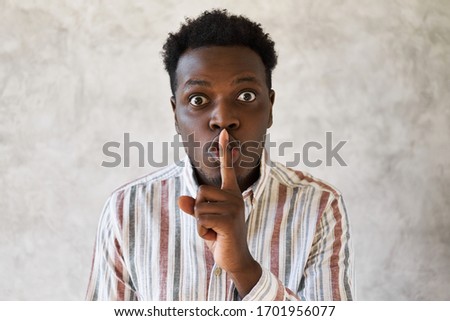 Excited emotional young African American male popping eyes out and putting finger on his lips, making Shush sound, asking to calm down or keep silent. Silence, secrecy and conspiracy concept