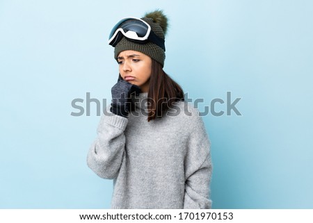 Mixed race skier girl with snowboarding glasses over isolated blue background having doubts.