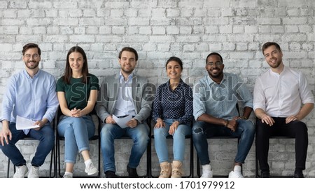 Portrait of smiling diverse multiracial young people sit in row on chairs wait for office interview, happy motivated multiethnic job candidates hired for vacant work position, employment concept Royalty-Free Stock Photo #1701979912