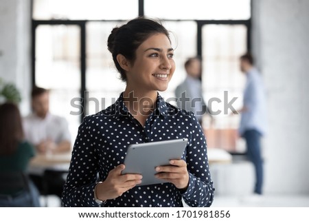 Smiling young Indian female employee hold tablet look in distance thinking, happy millennial biracial woman worker distracted from pad gadget, lost in thoughts visualizing, business vision concept Royalty-Free Stock Photo #1701981856