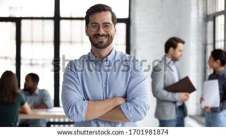 Smiling young Caucasian businessman in glasses stand in modern office show confidence and motivation at work, happy millennial European male CEO posing at workplace, leadership, success concept