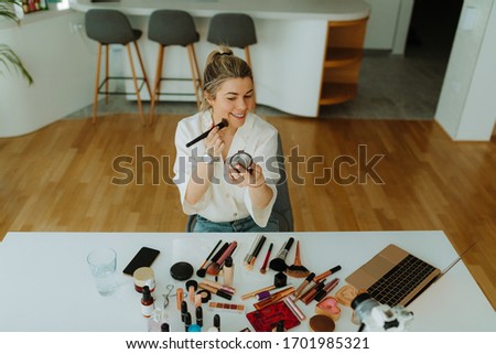 Photo of young smiling woman applying makeup with brush while recording her beauty blog about makeup and cosmetics at home.
