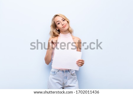 young blonde woman looking arrogant, successful, positive and proud, pointing to self holding a sheet of paper