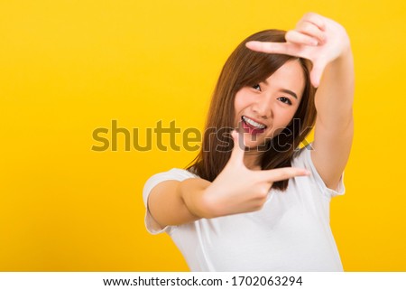 Asian happy portrait beautiful cute young woman standing wear t-shirt hands and finger making frame gesture camera for photography looking to camera, studio shot on yellow background with copy space
