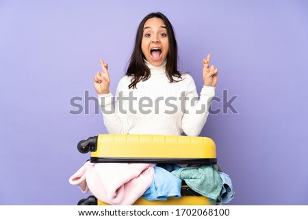 Traveler young woman with a suitcase full of clothes over isolated purple background with fingers crossing