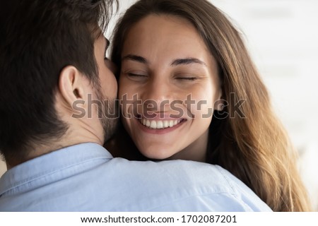 Close up head shot affectionate loving man cuddling beautiful young mixed race woman, whispering sweet words in ear. Caring husband showing love to bonding attractive wife, enjoying tender moment.