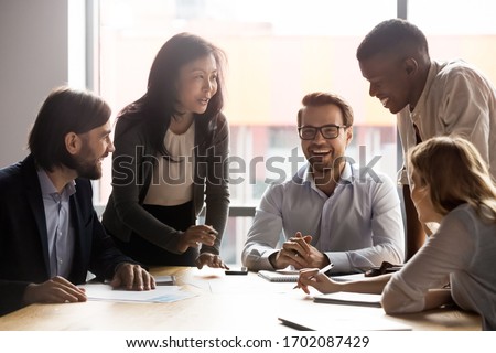 Smiling diverse businesspeople sit at office desk talk laugh discussing business ideas together, happy multiracial colleagues brainstorm joke negotiate at briefing in boardroom, cooperation concept Royalty-Free Stock Photo #1702087429