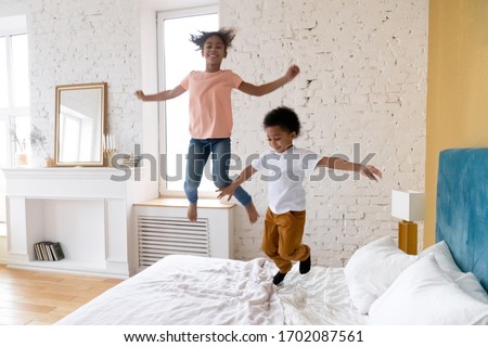 Happy african american children jumping at bedroom. Smiling and laugh diverse siblings enjoying weekend together at living room. Successful mortgage, cute kids on bed at new warm house. Royalty-Free Stock Photo #1702087561