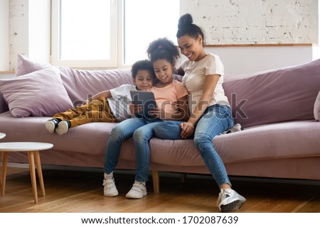 Happy african american mother looking at tablet screen with children. Diverse smiling family of mother, son and daughter sitting on couch using mobile device and gadget together.