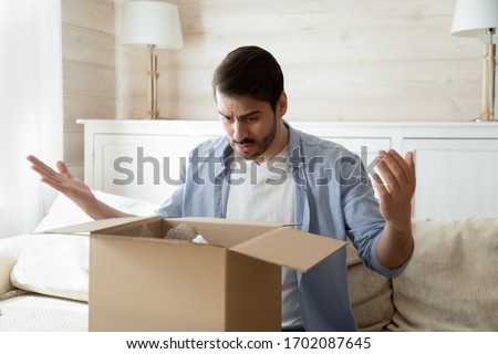 Unhappy young man looking inside delivery parcel, dissatisfied with damaged item. Outraged bearded guy sitting on couch in living room, feeling disappointed with crashed purchase from internet store. Royalty-Free Stock Photo #1702087645