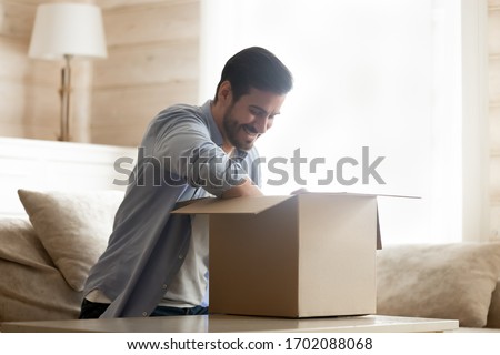 Smiling young man sitting on cozy couch at home, unpacking carton box with long-awaited purchased item. Happy male client satisfied with fast delivery service, unboxing order from internet store. Royalty-Free Stock Photo #1702088068
