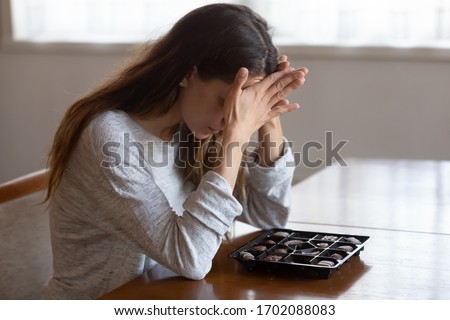 Depressed young mixed race woman sitting at table with chocolate sweets, feeling desperate alone at home. Addicted to candies stressed millennial girl suffering from eating disorder, anorexia concept. Royalty-Free Stock Photo #1702088083