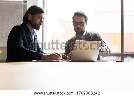 Serious middle-aged businessmen sit at office desk brainstorm discuss financial ideas using laptop, male business partners talk negotiate work on computer at meeting together, cooperation concept