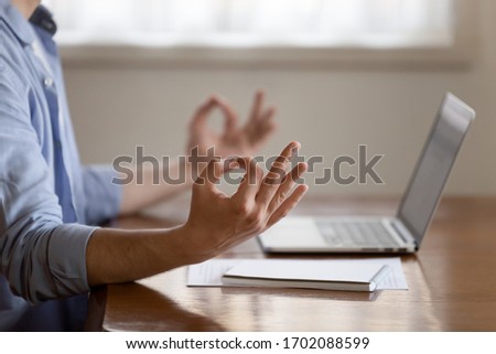 Close up focus on folded in mudra gesture male hands. Young peaceful man managing stress at workplace, meditating, calming down, doing yoga breathing exercises, sitting at table alone indoors. Royalty-Free Stock Photo #1702088599