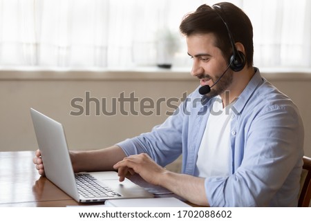 Side view young man wearing wireless headset with microphone, looking at laptop screen, study on online courses. Skilled male teacher giving online educational class to client, working remotely. Royalty-Free Stock Photo #1702088602