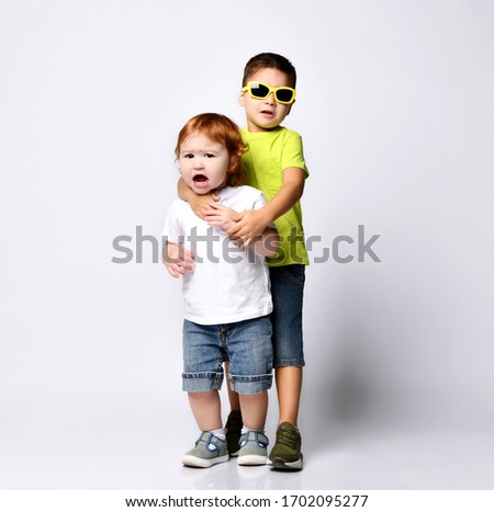 Brunet child in sunglasses is hugging by neck his little baby brother or sister who is screaming. Dressed in casual clothes, posing isolated on white background. Kids fashion. Full length, copy space