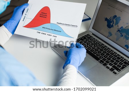 Doctor wearing protective gloves holding Flatten the Curve chart, sitting at the desk in front of laptop computer, Coronavirus COVID-19 global pandemic crisis protective measures to lower death toll  Royalty-Free Stock Photo #1702099021