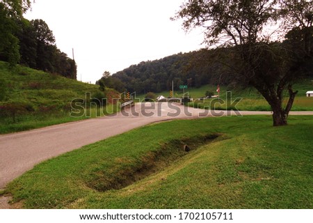 The ditch of a freshly cut yard in rural West Virginia, a forked road spanning off in different directions, a farmhouse and sloping ridges in the far distance.