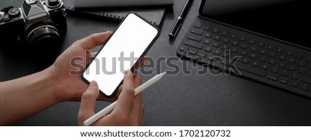 Cropped shot of male entrepreneur using mock-up smartphone while working with digital tablet, camera and other supplies on black table 