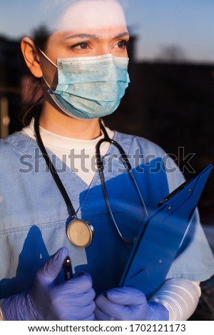 Serious female caucasian doctor looking through window with worried expression,lost hope due to high mortality rate death toll,Coronavirus COVID-19 pandemic crisis,overworked exhausted UK EMS staff  Royalty-Free Stock Photo #1702121173
