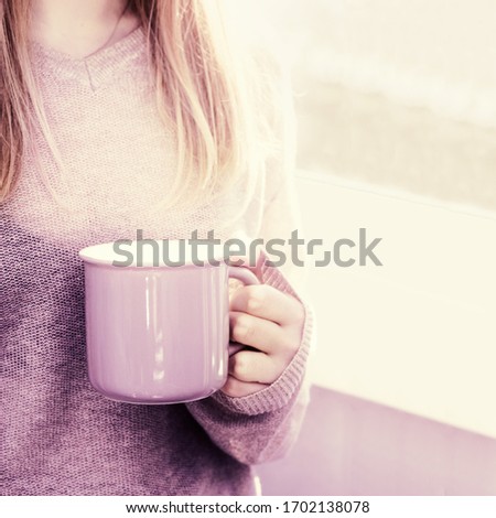 Beautiful blonde woman holding big cup of tea  and stands at window. Good morning and enjoy the sunshine. Domestic life and happy concept. Toned image.