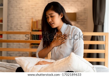 Portrait of 20s young Asian woman having difficulty breathing in bedroom at night. Shortness of breath, asthma, difficult to breathe problems. Corona Virus symptoms. Royalty-Free Stock Photo #1702189633