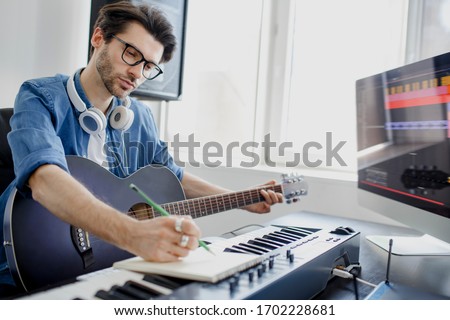 Male music arranger composing song on midi piano and audio equipment in digital recording studio. Man plays guitar and produce electronic soundtrack or track in project at home. Royalty-Free Stock Photo #1702228681