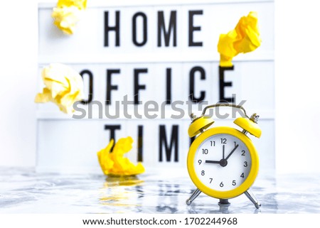 lightbox with text HOME OFFICE TIME in front concrete background with alarm clock, copy space, banner for freelance coronavirus covid-19 quarantine flying crumpled yellow papers, business planning and