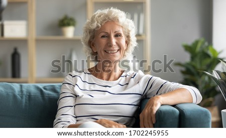 Head shot portrait smiling beautiful mature woman sitting on couch at home, looking at camera, happy excited older female with curly grey hair resting on cozy sofa, posing for photo in living room