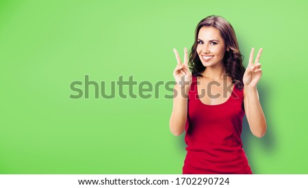Young happy smiling woman in casual clothing, showing two fingers or victory gesture, over green color background. Happy girl in red dress. Brunette excited model.