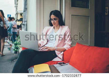 Hispanic woman in classic eyewear searching information during freelance job with web content and graphic design, skilled female watching webinar video connecting to wifi internet in sidewalk