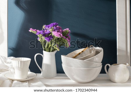 A bouquet of limonium and a set of white dishes: porcelain milk jug, coffee cup, bowls on a white rack on a dark blue background. Kitchen