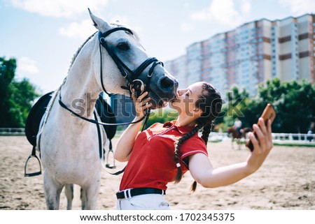 Caucasian female jockey kissing thoroughbred mare in paddock country club and clicking selfie pictures for sharing media content to social networks, millennial making photos with friendly stallion