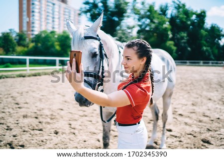 Caucasian female jockey enjoying free time for horseback in paddock country club and clicking pictures for sharing media content to social networks, millennial making selfie vlog with friendly mare