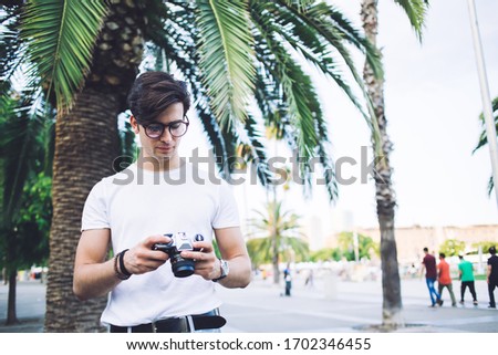 Pensive caucasian male in eyewear holding camera checking photos while explore town on vacation trip, skilled man photographer standing on city square with palm trees enjoying summer weekends