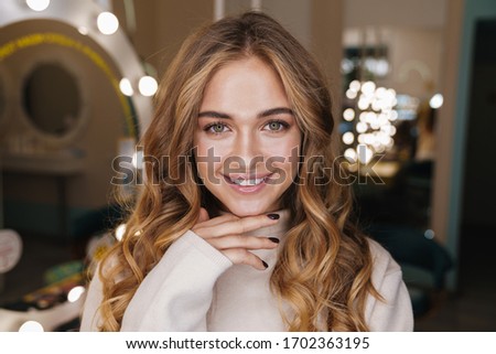 Photo of a young happy optimistic cute cheery blonde girl indoors in beauty salon looking at camera.